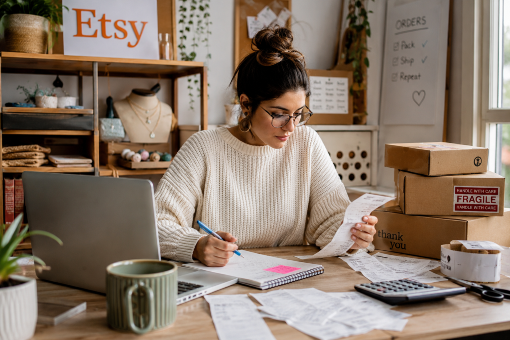 An Etsy seller working at a tidy desk with handmade product samples, shipping boxes, receipts, a laptop, and a calculator, organizing inventory and tracking business finances in a warm, well-lit home workspace.